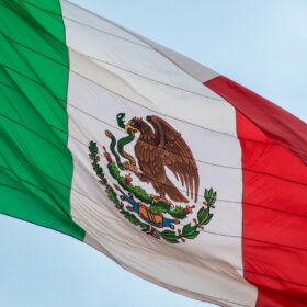 The Mexican flag with its eagle emblem waving against a blue sky during Hispanic Heritage Month